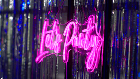 Close-Up-Of-Neon-"Let's-Party"-Sign-Hanging-Against-Tinsel-In-Nightclub-Or-Funfair